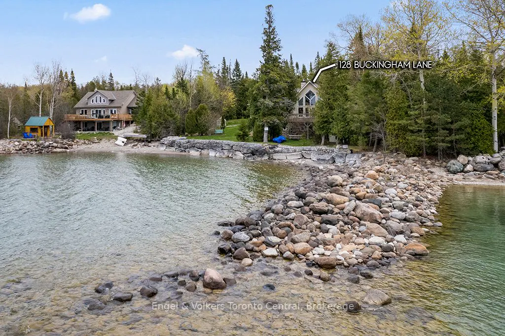 Waterfront view of 128 Buckingham Lane, a house nestled among trees. A rocky shoreline leads to the water. Another house is visible in the background.