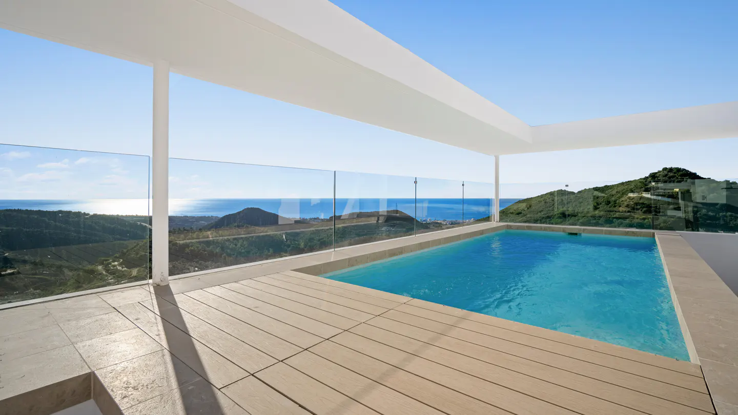 Outdoor pool with wood deck and glass railing overlooking ocean and green hills under a blue sky.