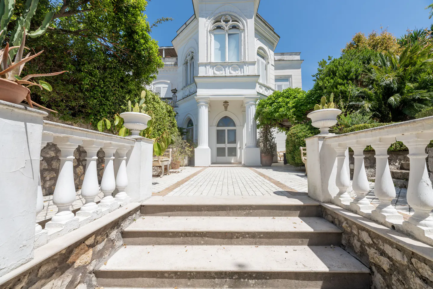 Exterior view of a white villa with stone steps, balustrades, and lush greenery under a clear blue sky.