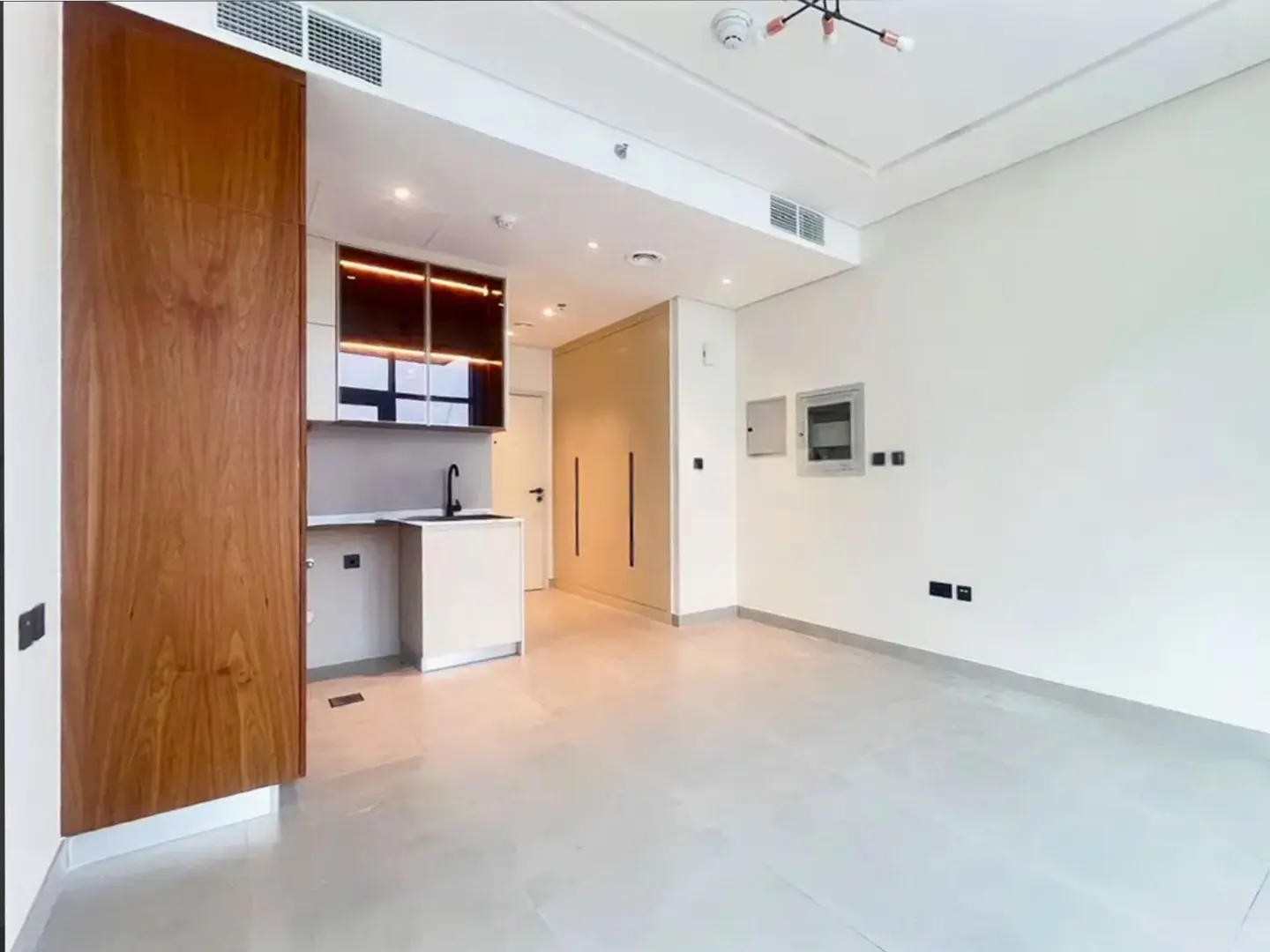 A bright, empty studio apartment with white walls, light gray tile floors, and a small kitchen area with wood cabinets.