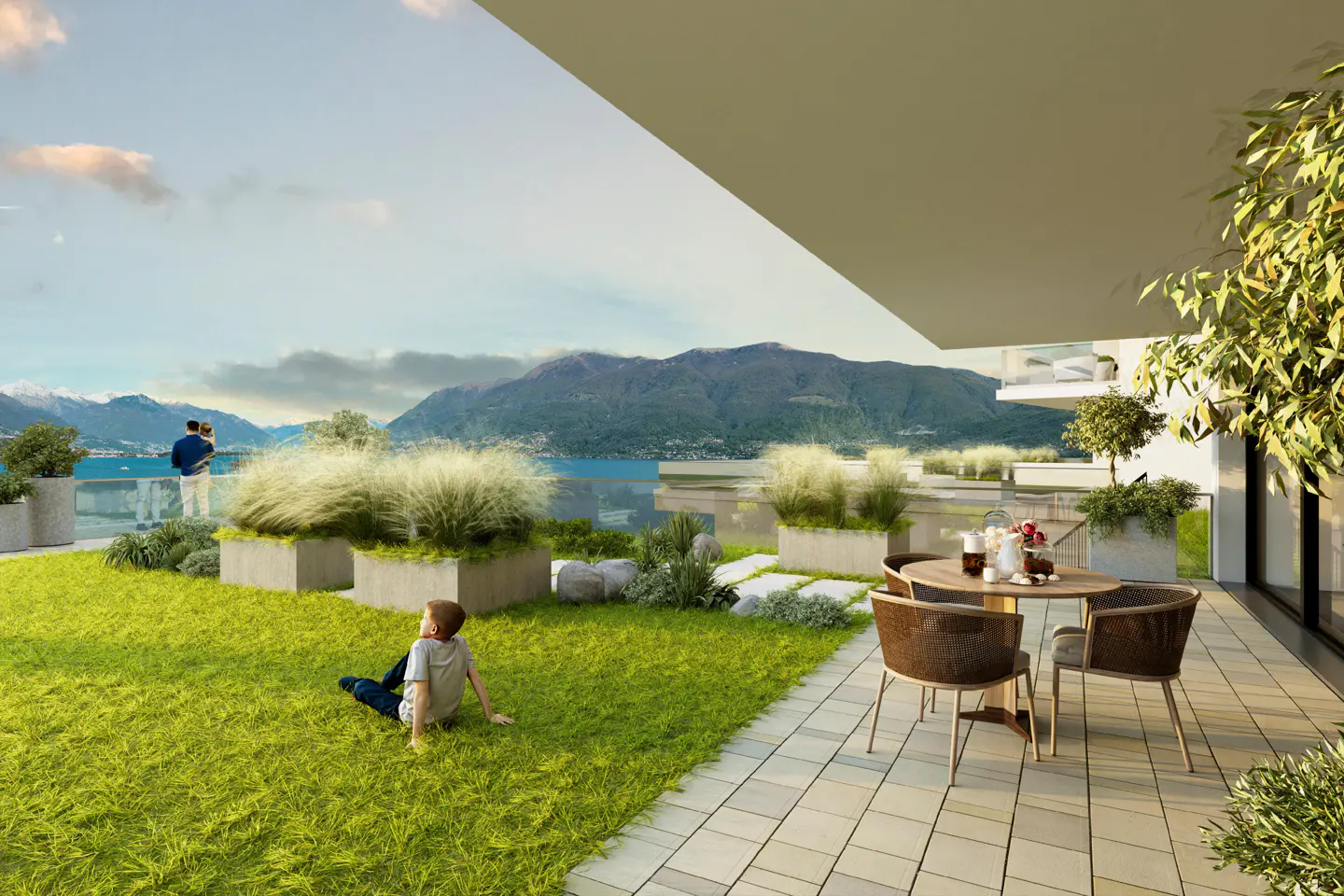 A child sits on a grassy terrace overlooking a lake and mountains, with a patio table and chairs nearby.