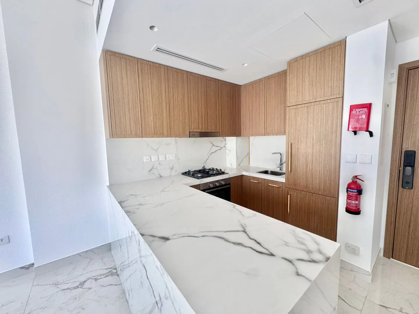 A modern kitchen with white marble countertops and light wood cabinets. A red fire extinguisher is mounted on the wall.