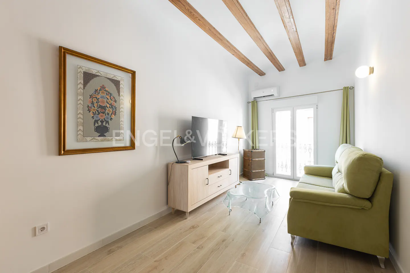 Bright living room with wood floors, white walls, and exposed beams. A green sofa faces a TV on a light wood cabinet. Artwork hangs on the wall.