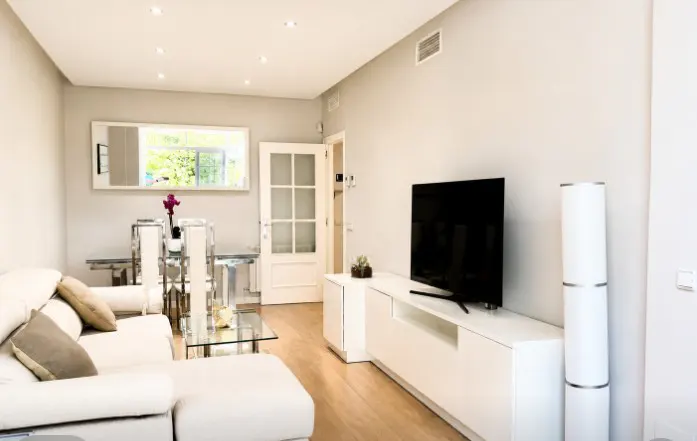Modern living room with a white sectional sofa, glass coffee table, TV on a white console, and a dining area in the back.