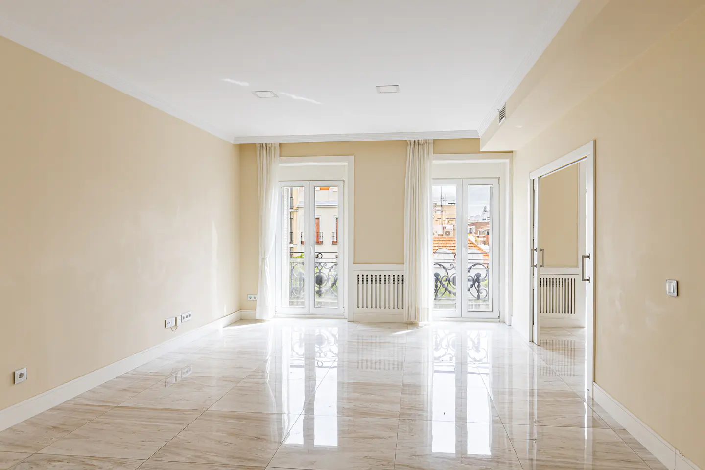 Bright, empty room with beige walls, marble floors, and two sets of French doors with white curtains. A doorway is visible on the right.