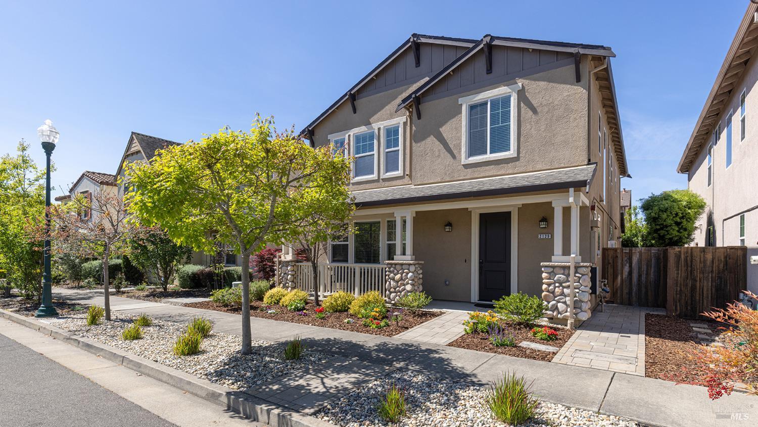 Two-story tan house with a black door, white trim, and stone accents on the porch columns. A green tree is in the front yard.