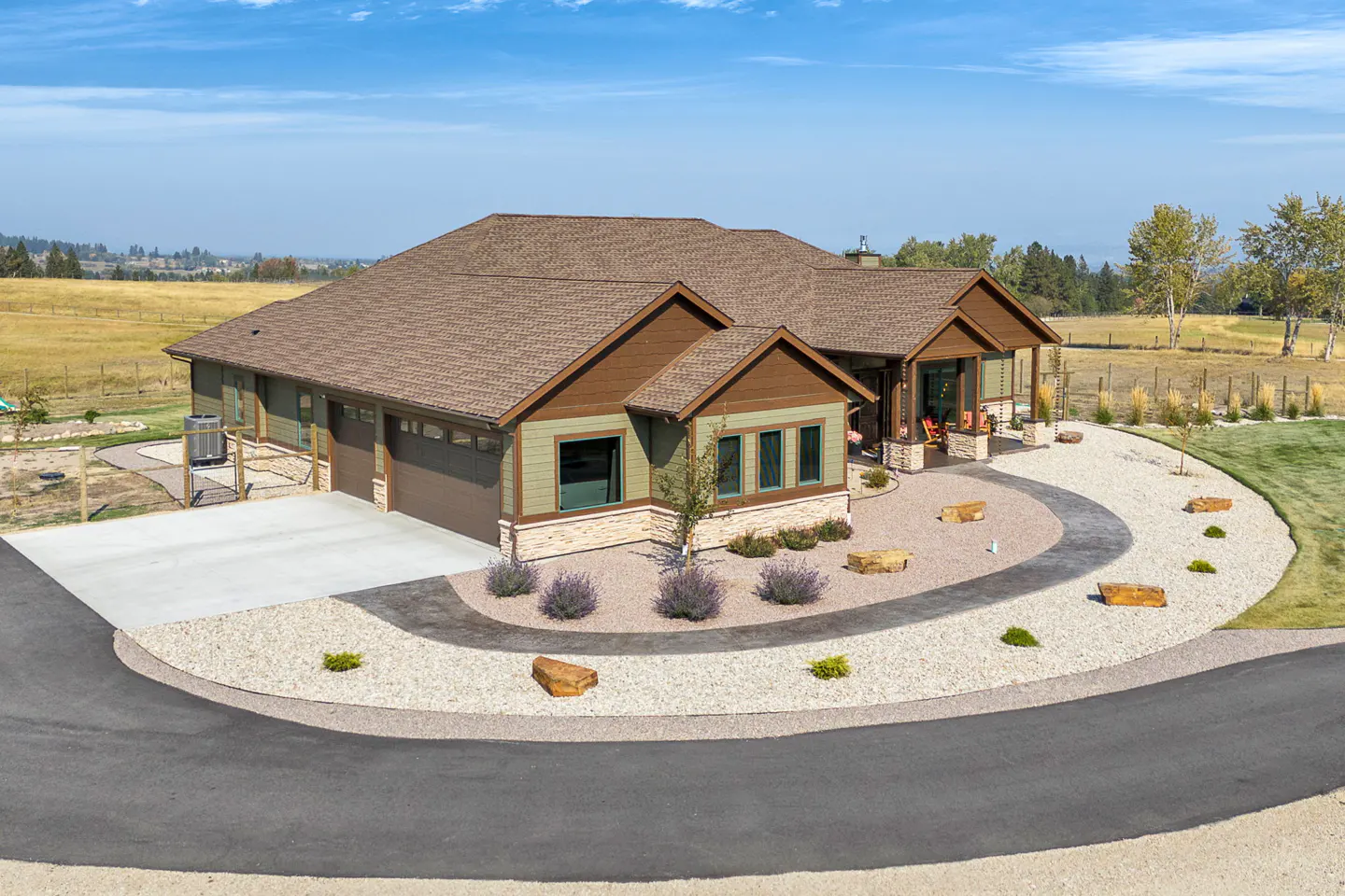 Aerial view of a green and brown single-story house with a brown roof, surrounded by a gravel driveway and grassy lawn.