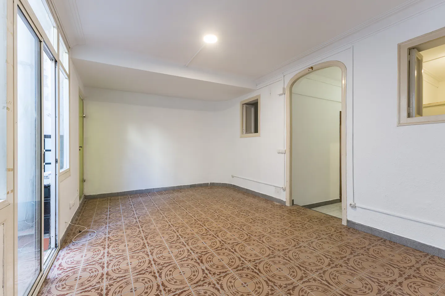 Empty room with white walls, brown patterned tile floor, arched doorway, and sliding glass doors to the outside.