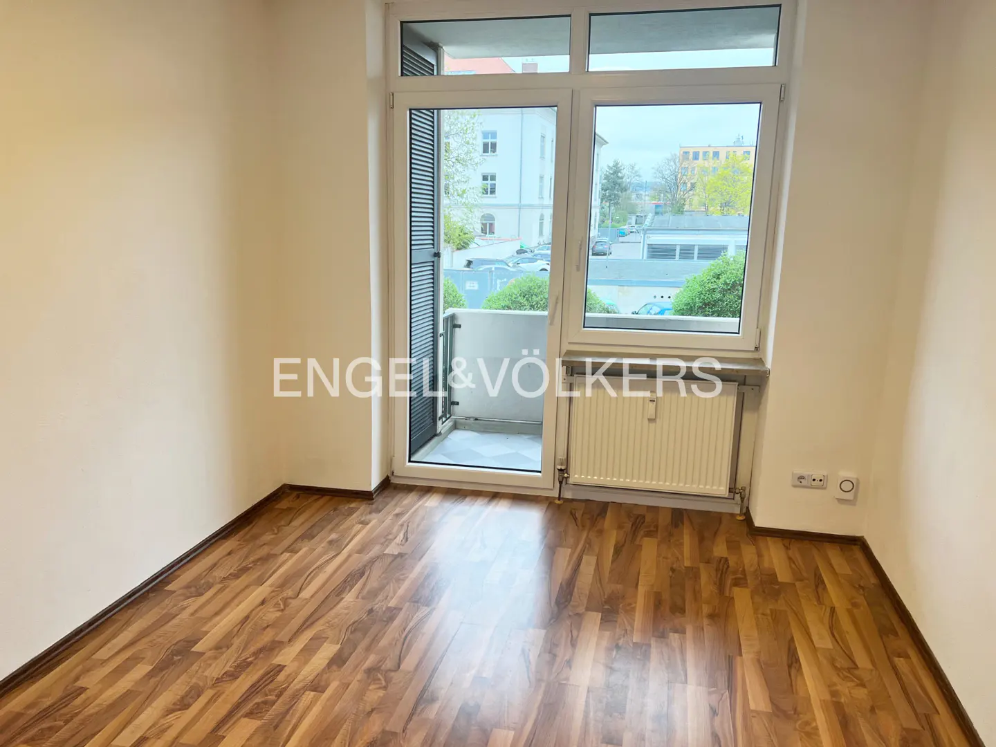 Empty room with wood floors, white walls, and a balcony. A radiator sits under the window. Engel & Völkers logo is visible.
