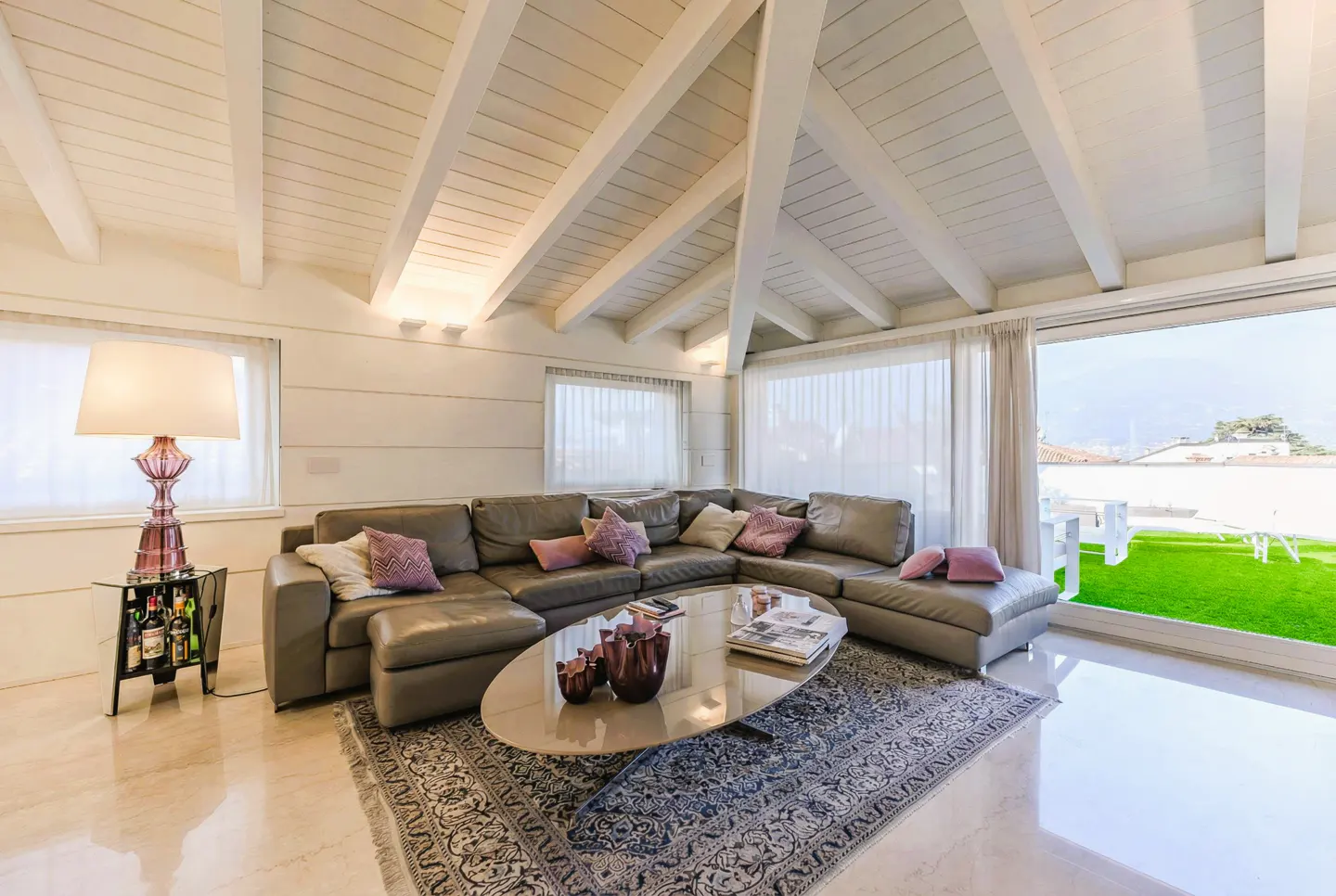 Living room with white vaulted ceiling, gray sectional sofa, oval coffee table, and large window overlooking a green lawn.
