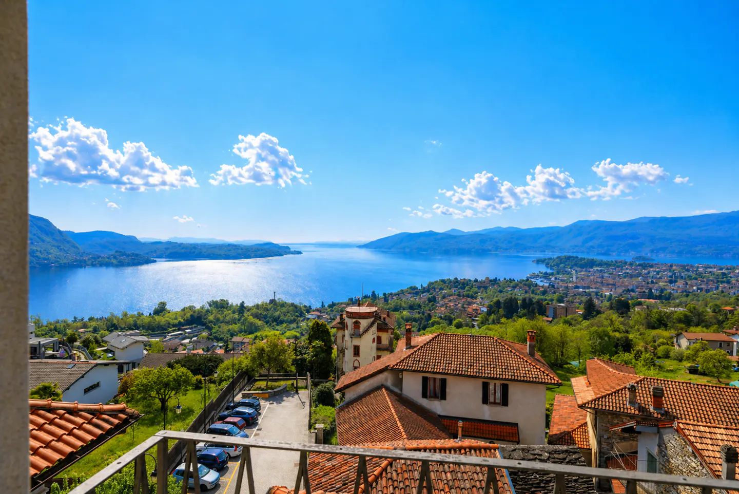 View from a balcony overlooking Lake Maggiore, Italy, with red-tiled roofs, green trees, and a bright blue sky.