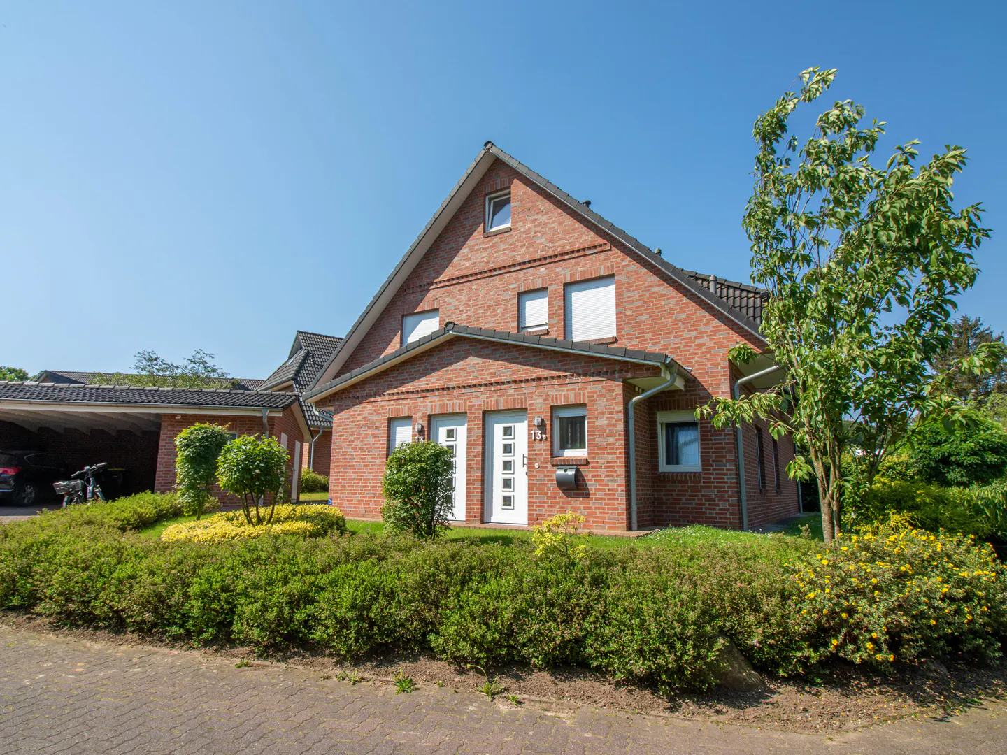 Two-story brick house with white doors and windows, green bushes, and a carport under a clear blue sky.