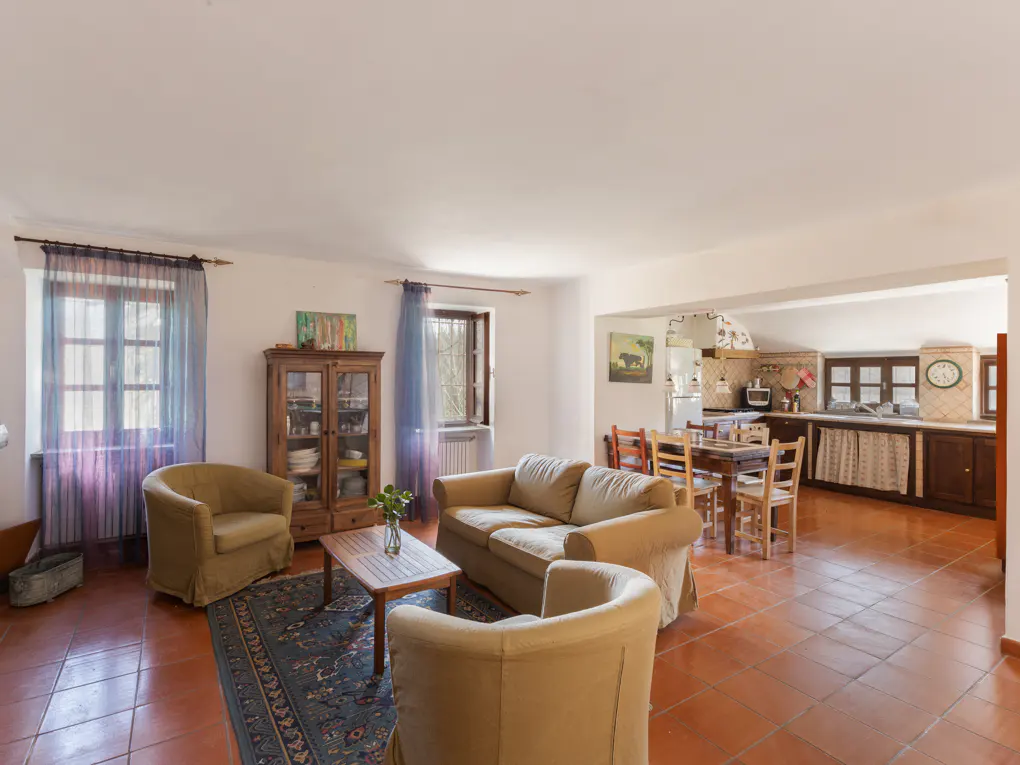 A bright living room with terracotta tile floors, tan sofas, a wooden cabinet, and an open kitchen in the background.