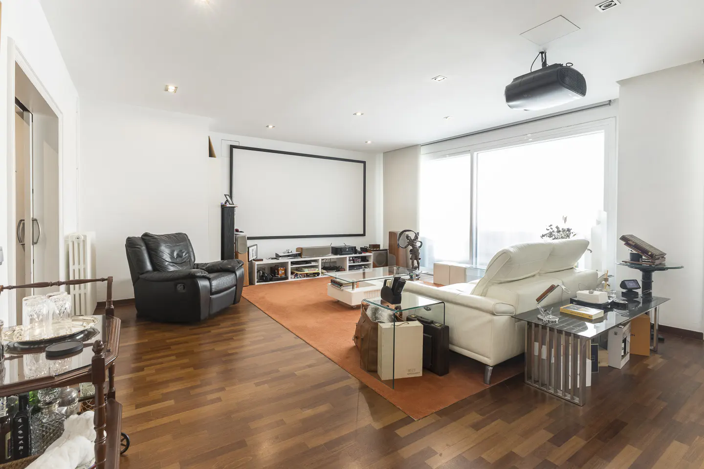 Bright living room with hardwood floors, white walls, and a large projector screen. A black leather recliner and white sofa provide seating.