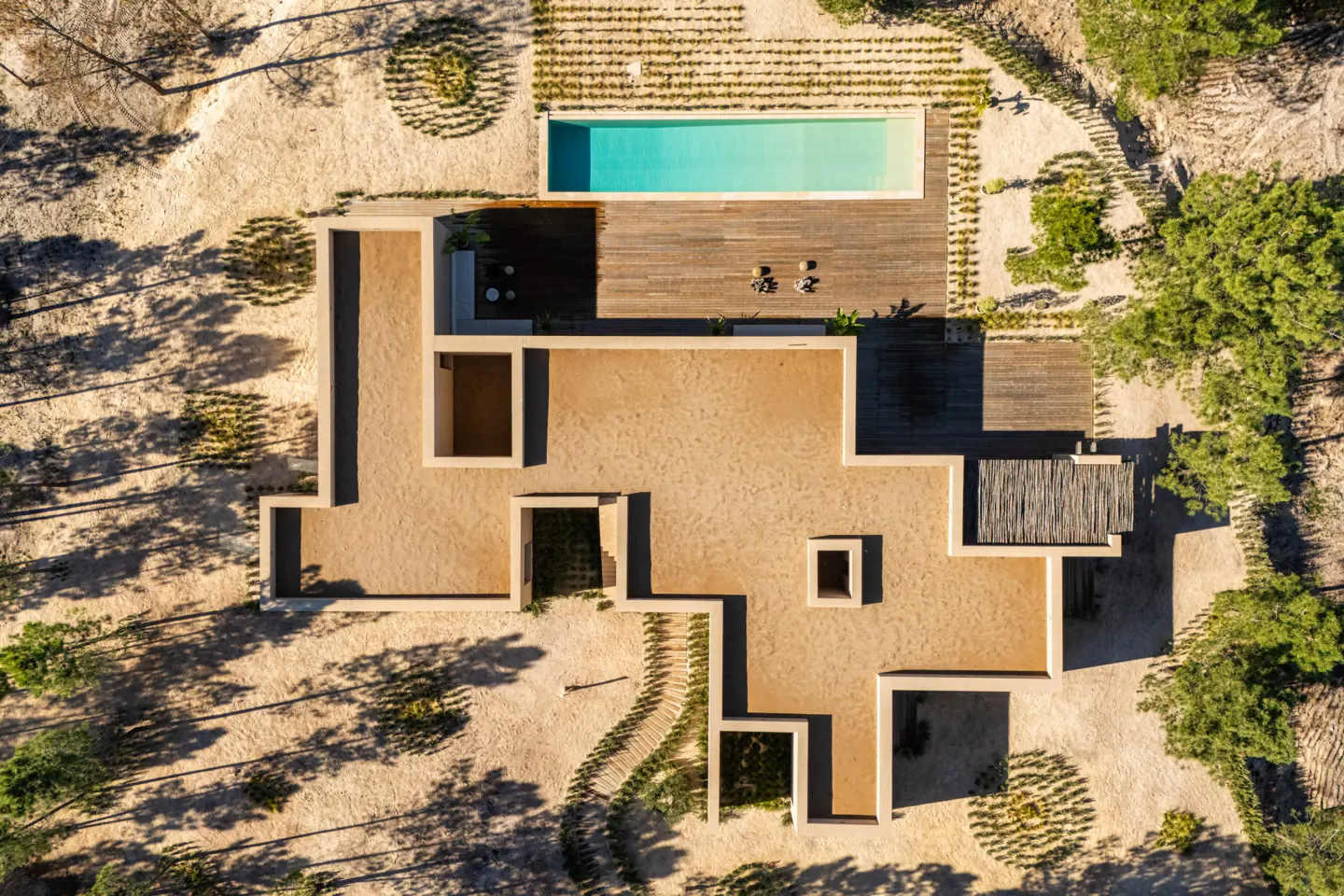 Aerial view of a modern, tan-colored house with a pool and wooden deck surrounded by trees and sand.