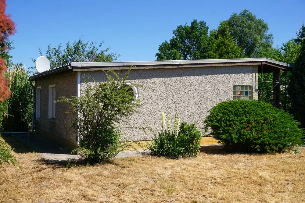 A one-story beige house with a satellite dish on the roof, surrounded by green bushes and dry grass.