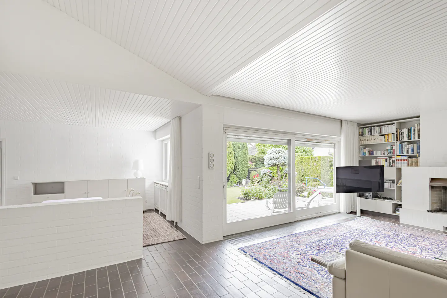 Bright, modern living room with white paneled ceilings, tiled floors, a large patterned rug, and a view of the garden.