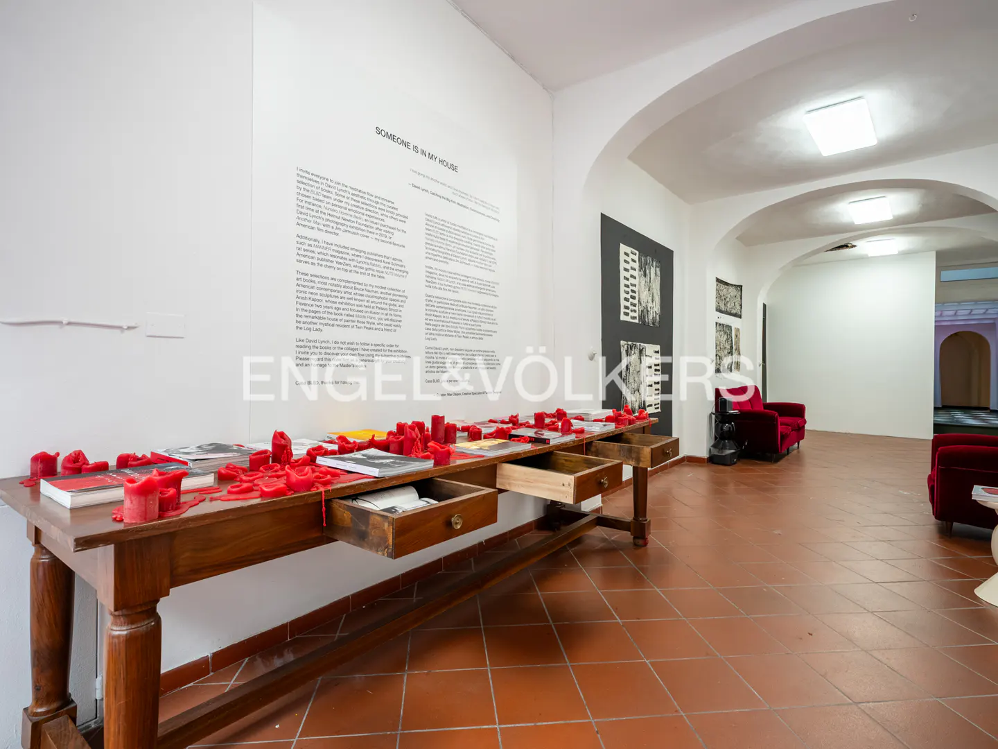 A long wooden table covered in red wax drips and books sits before a wall with text, inside a gallery with red seating.
