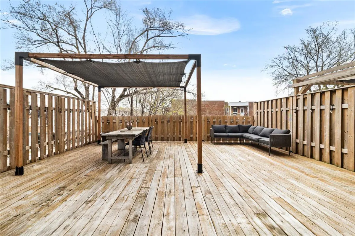 Wide shot of a wooden deck with a dining table under a pergola and a gray sectional sofa. A wooden fence surrounds the deck.