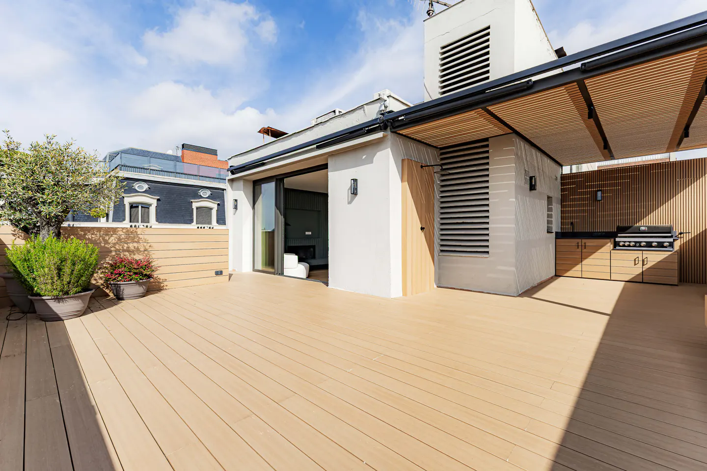 Wide shot of a rooftop deck with a grill, outdoor kitchen, and sliding glass doors to the interior. The deck is made of light brown wood.