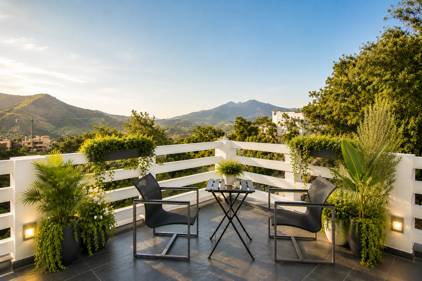 Outdoor patio with two black chairs and a small table, overlooking green hills and mountains under a blue sky. White railing with plants.