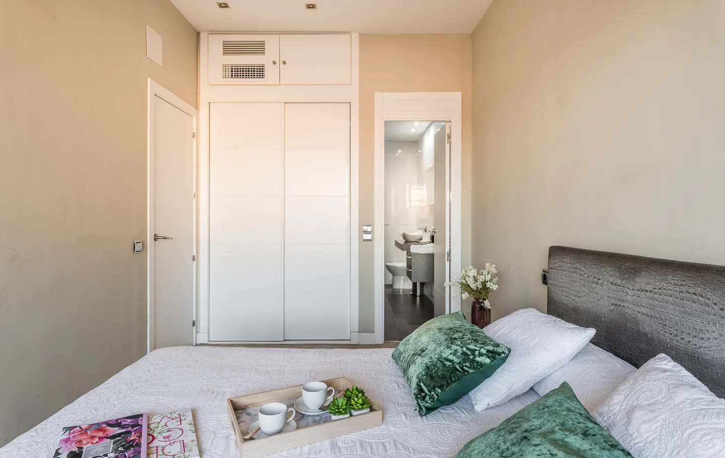 Bedroom with a white bed, green pillows, and a tray with cups. A bathroom is visible through an open door.