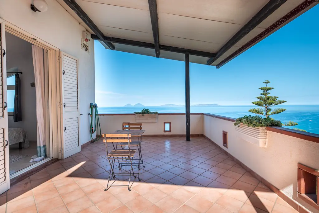 Covered terrace with tiled floor, small wooden table and chairs, overlooking the blue sea and distant islands.
