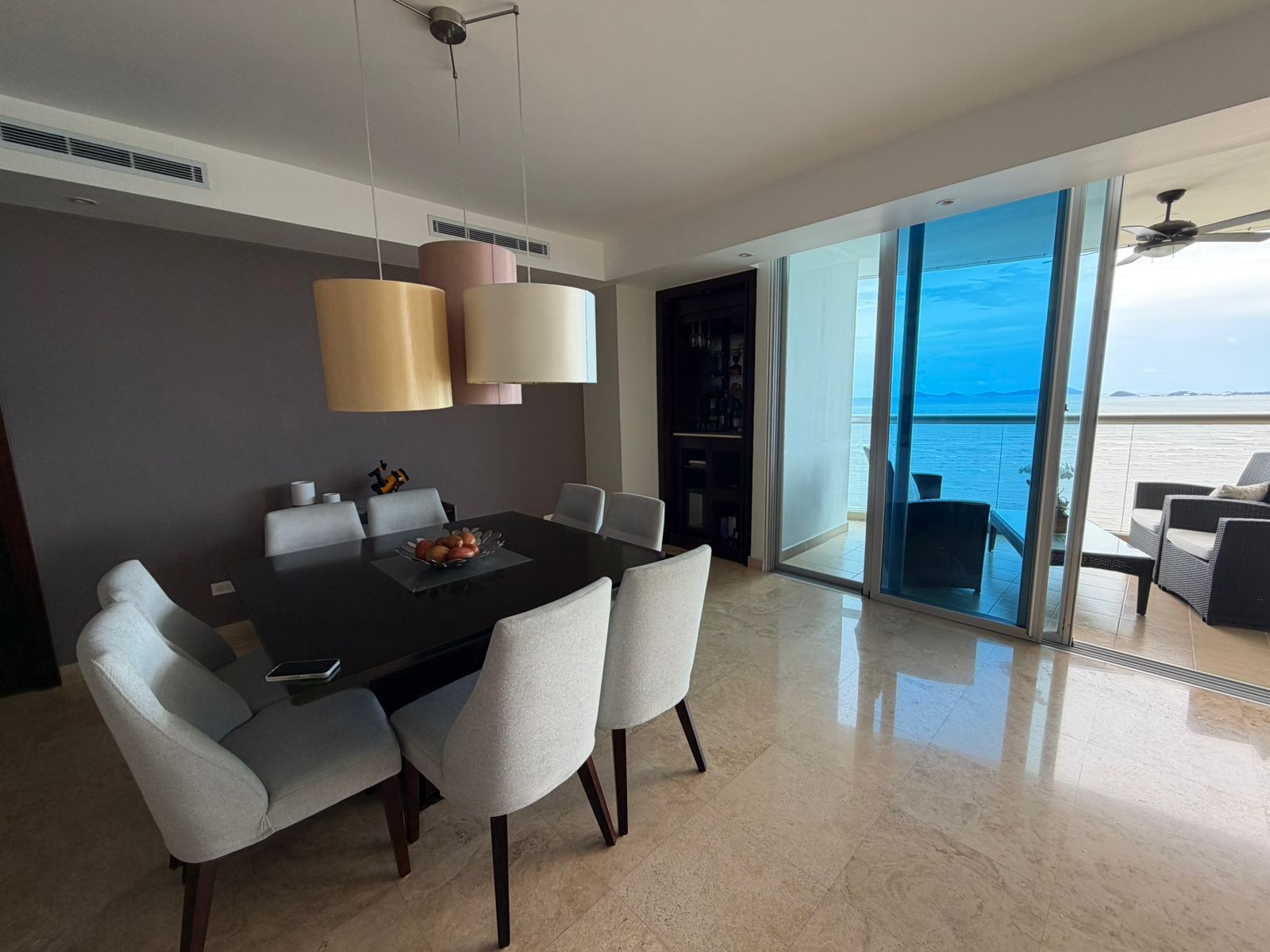 Modern dining room with a dark table, light gray chairs, and pendant lights, opening to a balcony with ocean views.