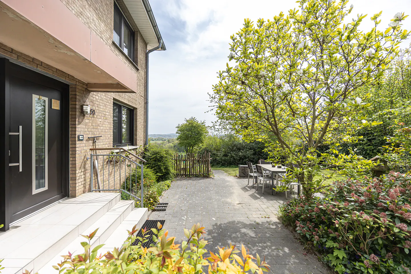 Brick house with a black front door and white steps leading to a stone patio with a table and chairs. A large tree with white flowers is on the right.