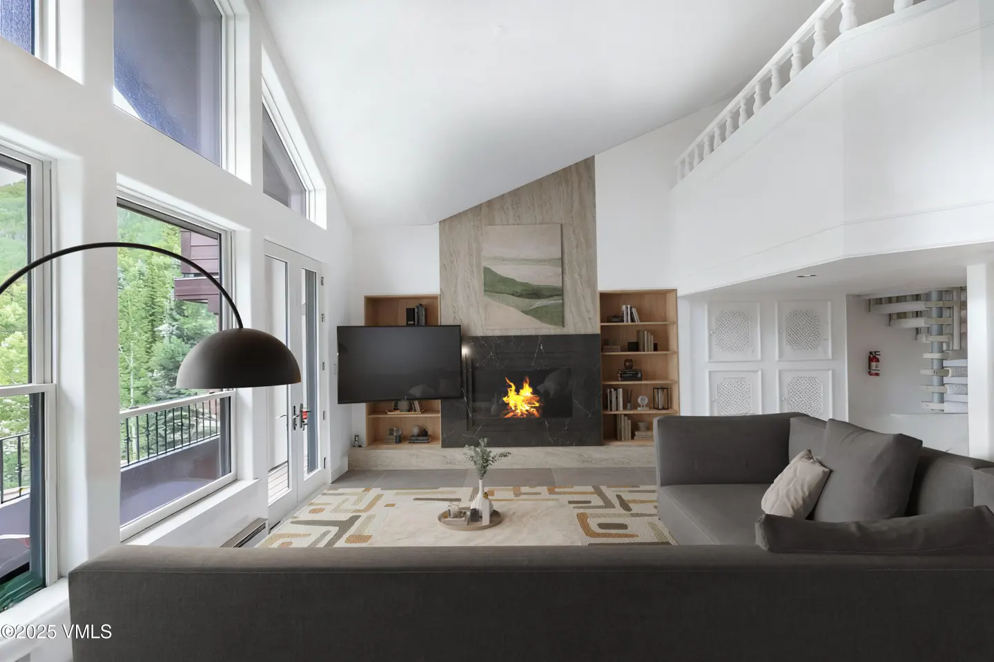 Bright living room with a gray sectional sofa, fireplace, TV, and large windows with a view of trees.