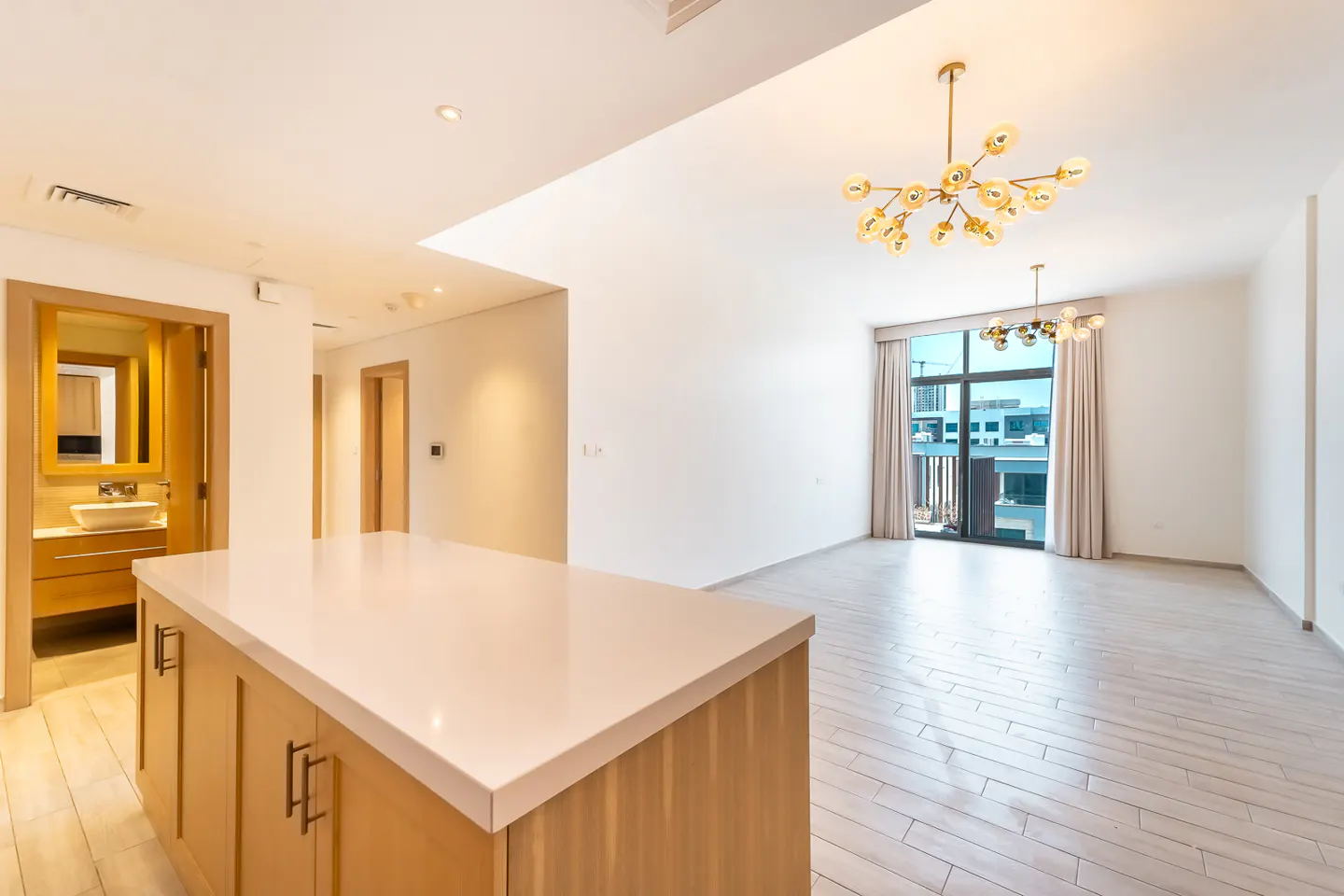 Bright, modern apartment interior with a kitchen island, wood floors, and a large window with city views. A gold chandelier hangs above.