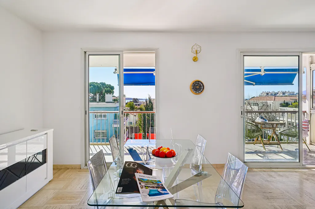 Bright dining room with glass table, clear chairs, and magazines. Balconies with blue awnings offer city views. White walls, clock, and modern cabinet.