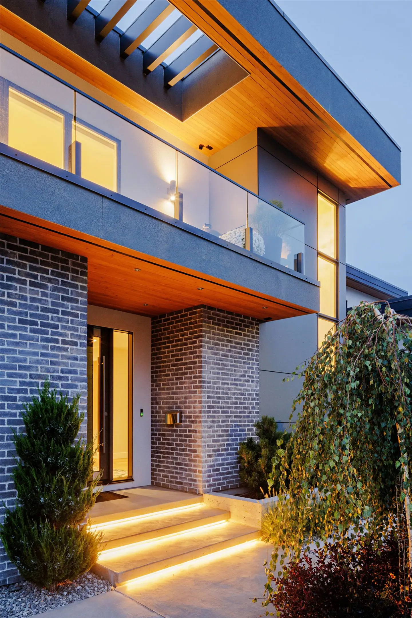 Modern home exterior with brick accents, illuminated steps, and a glass-railed balcony under a wooden pergola.