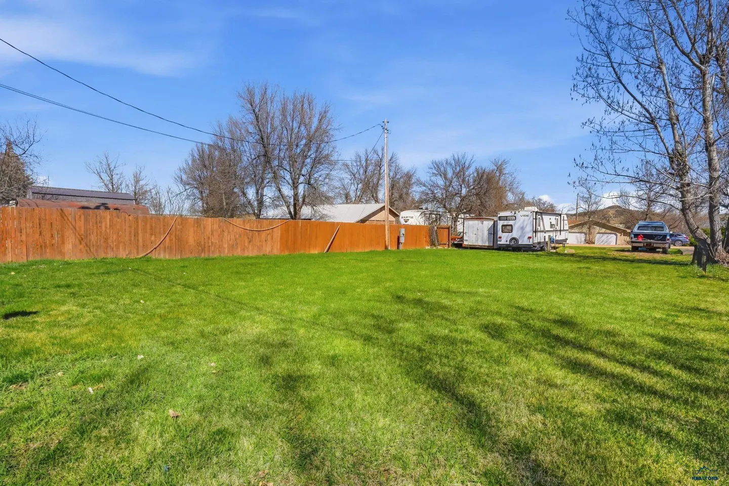 A grassy lot with a wooden fence, RVs, and a car under a blue sky.