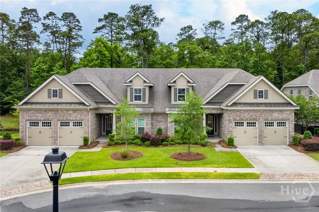Two-story brick duplex with tan garage doors, dormer windows, and a gray roof. Green lawn and trees in front.