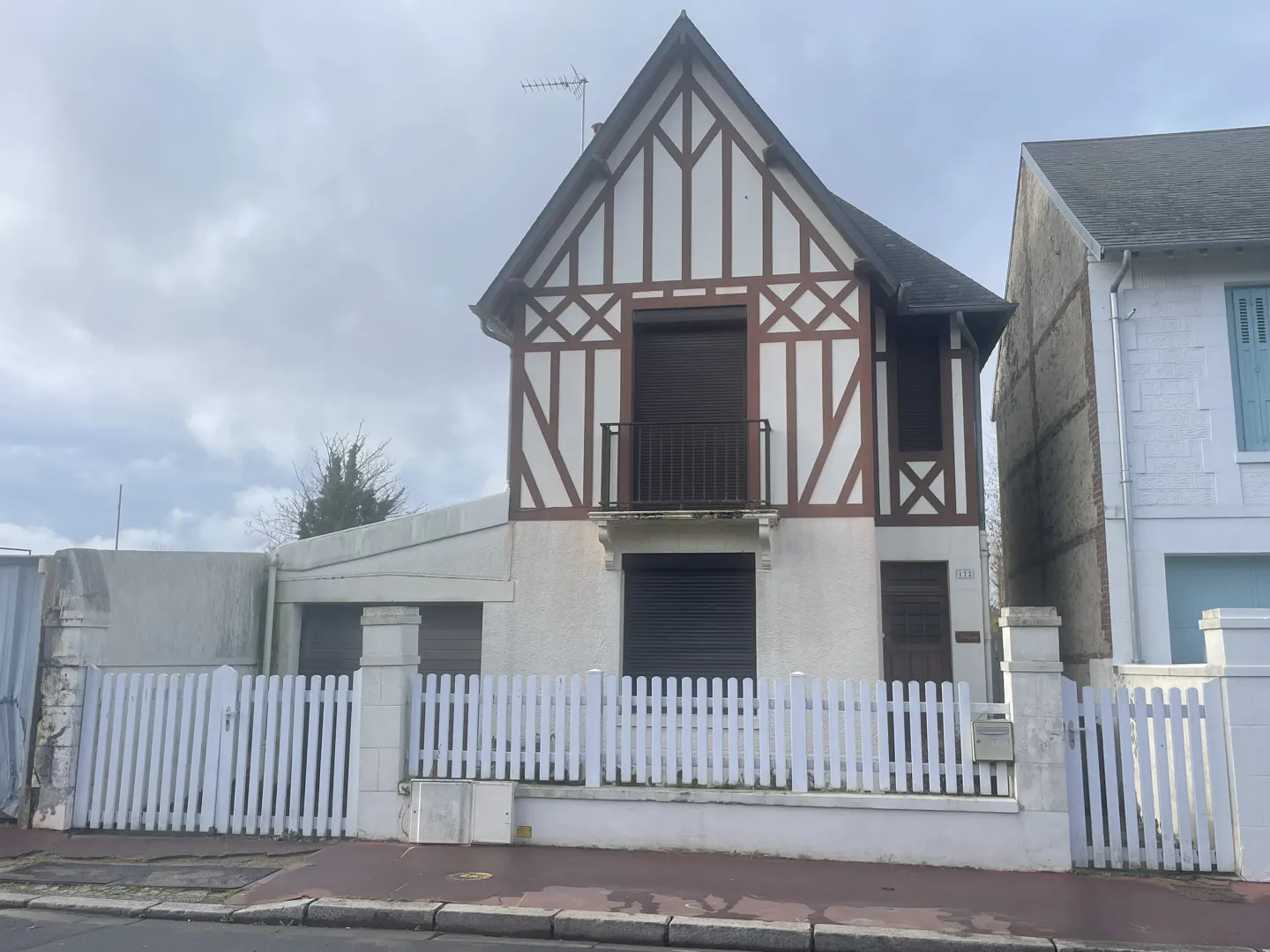 Two-story house with white and brown Tudor-style facade, black roof, and white picket fence on a cloudy day.