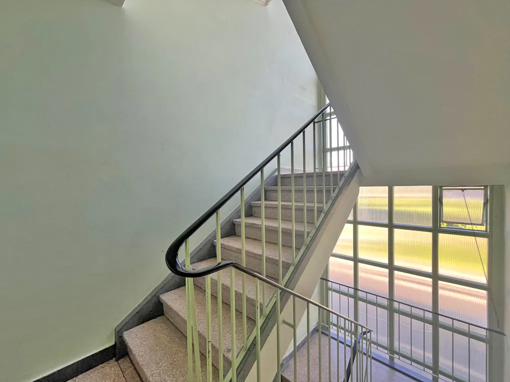 A stairwell with stone steps, light green walls, and a black handrail. Windows with metal frames are on the right.