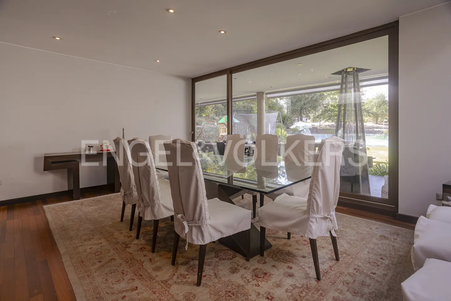 Dining room with a glass table, chairs with white covers, and a large window overlooking a pool and garden.
