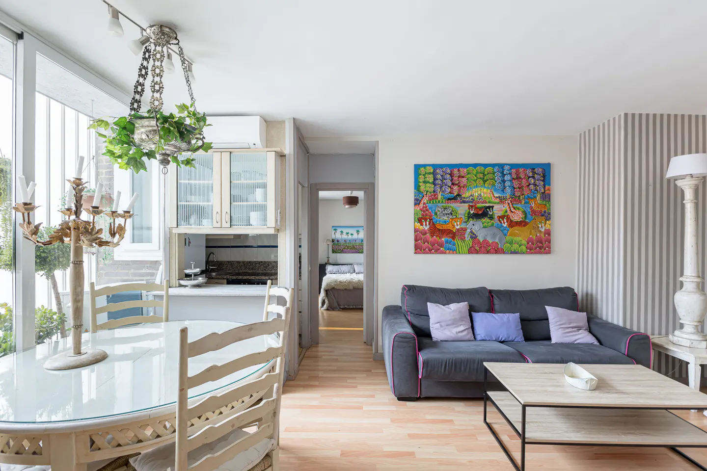 Bright living space with a gray sofa, colorful art, and a dining area with a glass table and white chairs. A bedroom is visible through a doorway.