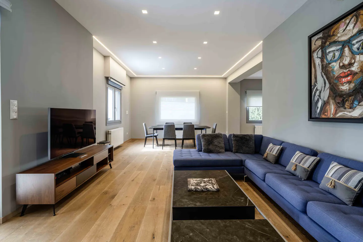 Modern living room with a blue sectional sofa, dark coffee table, wooden media console, and dining area on hardwood floors.