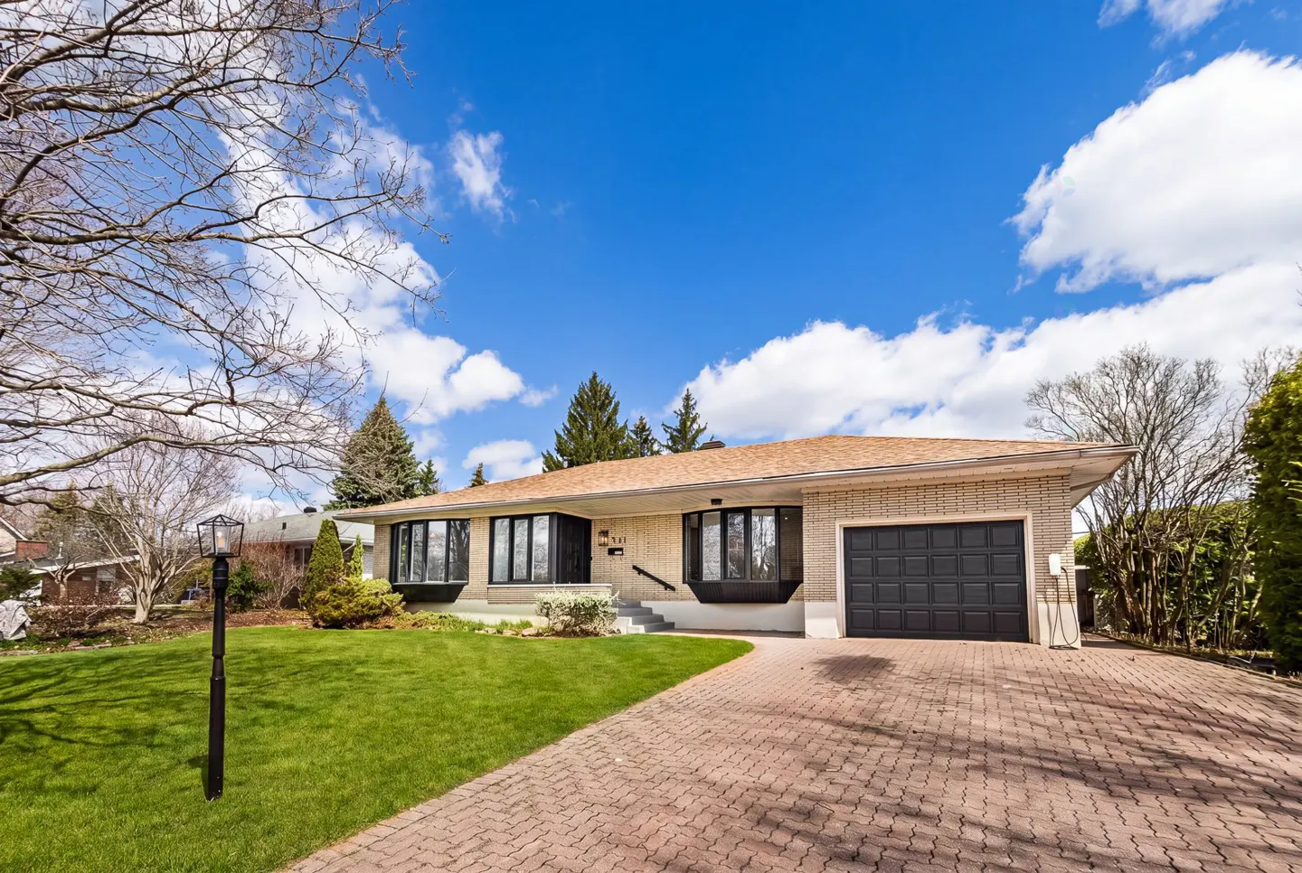 Tan brick ranch-style house with a black garage door, bay windows, and a brick driveway under a blue sky.