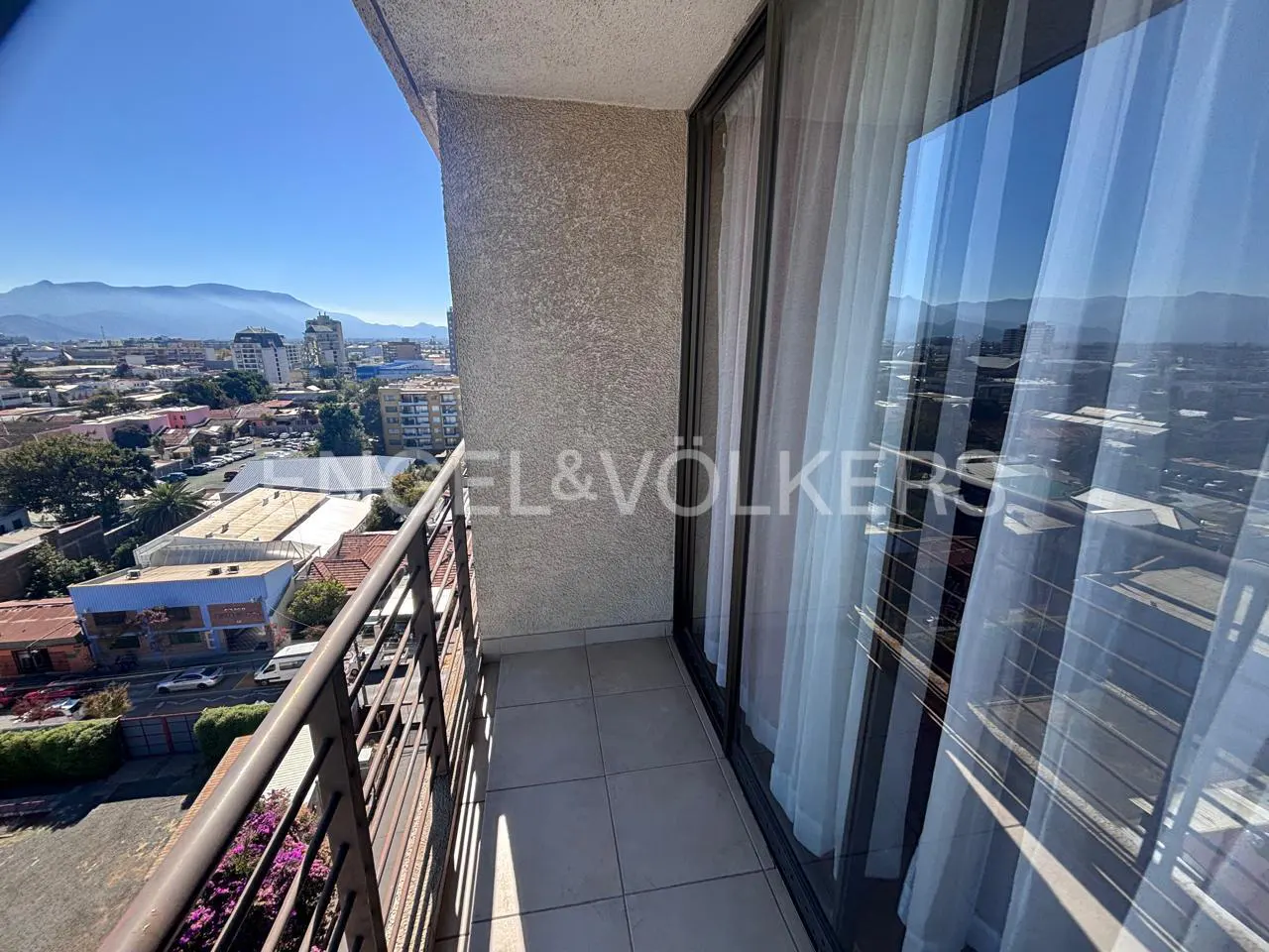 Balcony view with metal railing, tile floor, and sliding glass doors with white curtains. Cityscape and mountains visible in the background.