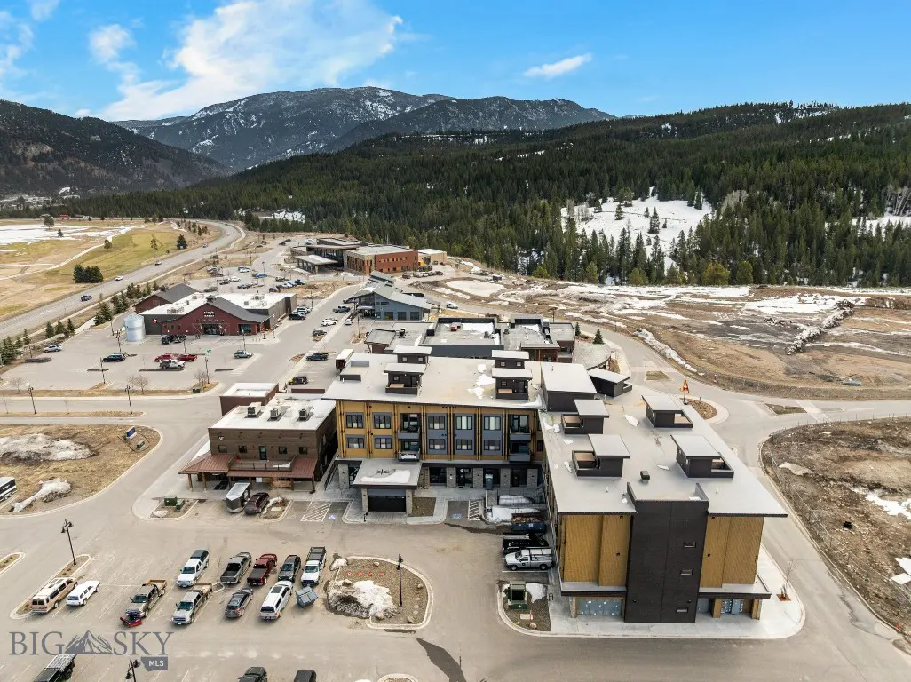 Aerial view of a multi-story building with a parking lot, surrounded by mountains and trees under a blue sky.
