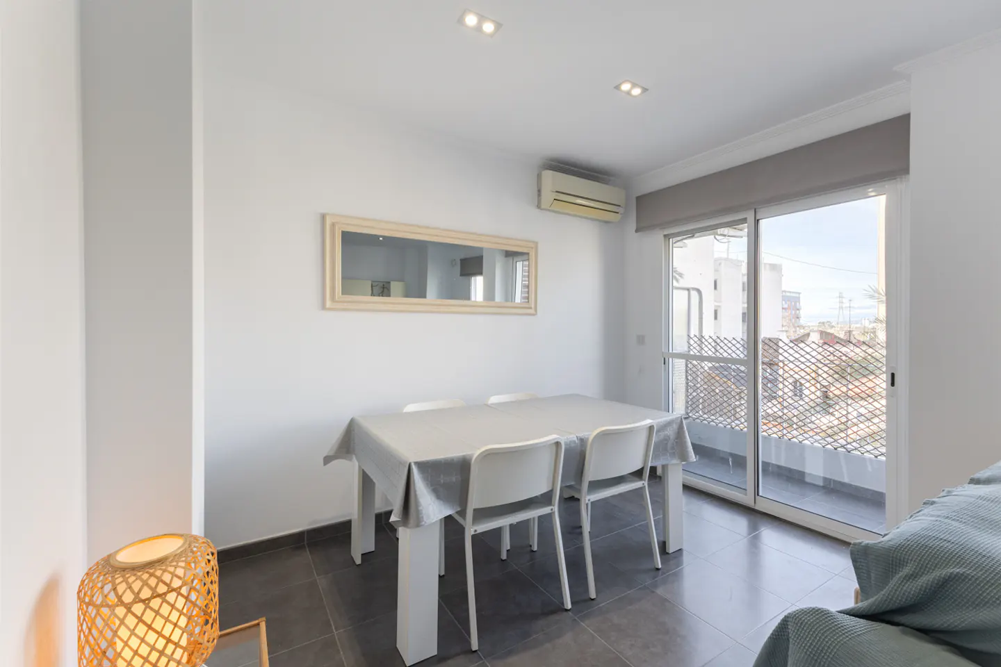 Bright dining room with white walls, gray tile floor, and a white table with four chairs. A mirror hangs above the table, reflecting the room. Sliding glass doors lead to a balcony.