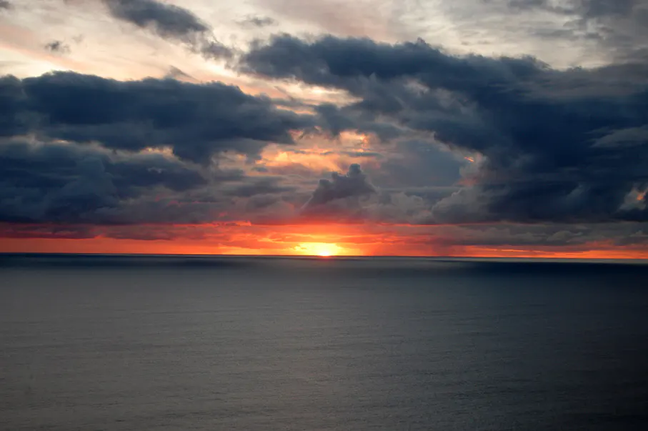 Ocean view at sunset with dark clouds above. The sun is setting on the horizon, creating a red glow.
