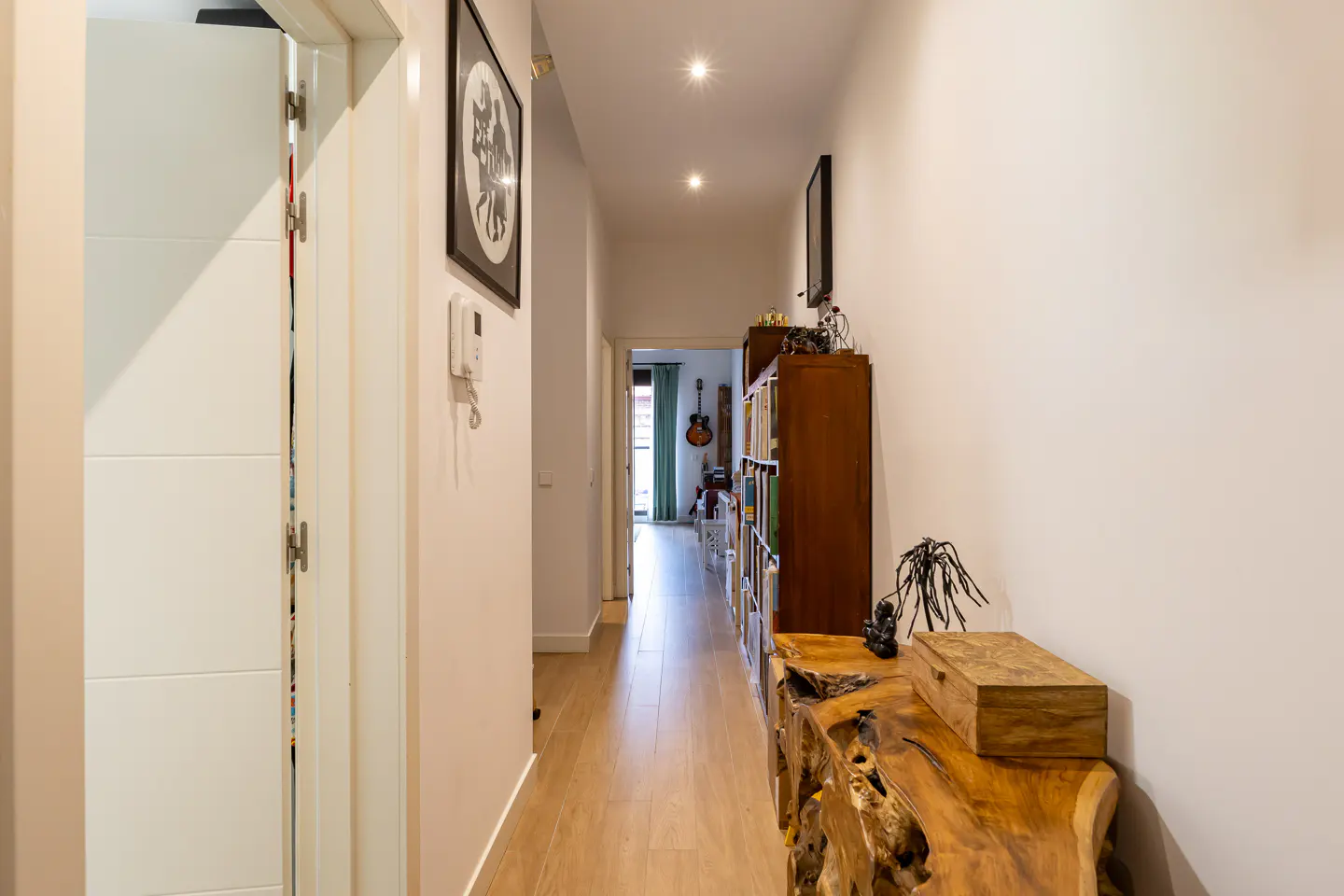 Hallway with light wood floors, white walls, and recessed lighting. A wooden console table and bookcase line the right wall. Artwork hangs on the left.