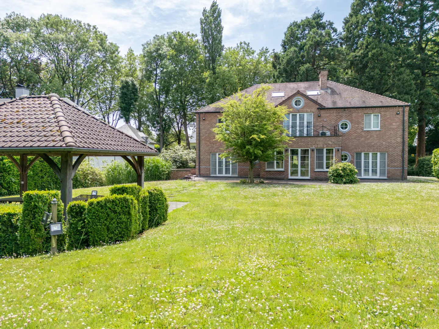 Brick house with a gazebo on a green lawn. The house has a balcony and round windows. Trees surround the property.