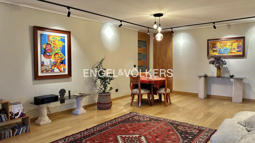 A living room with hardwood floors, a red patterned rug, and track lighting. There is a round table with four chairs, and two paintings on the walls.
