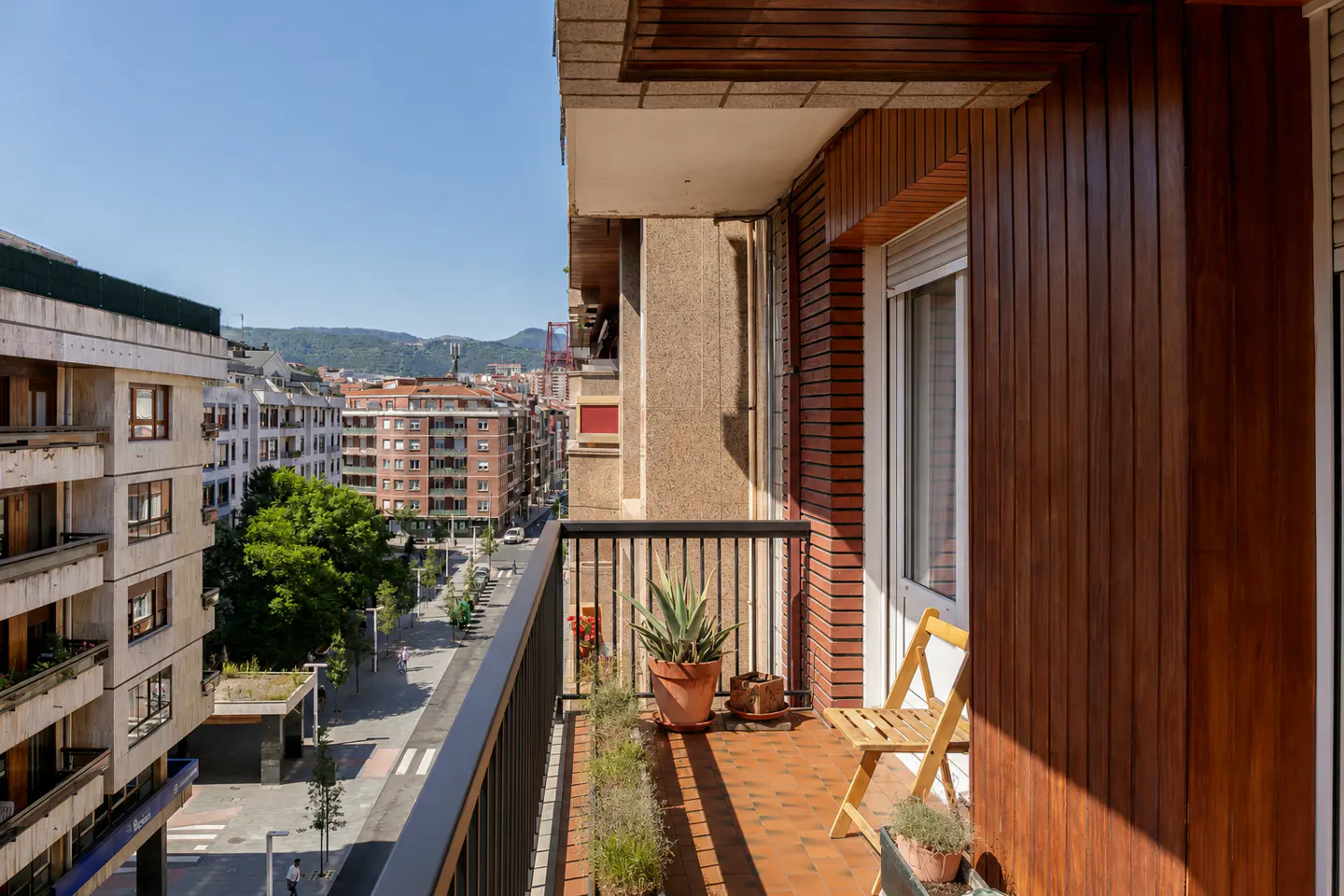Balcony view of a city street. Potted plants line the railing, with a wooden chair on the brick floor. Buildings and mountains are visible in the background.
