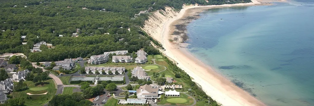 Aerial view of a coastal resort with a sandy beach, blue ocean, green forest, and residential buildings.